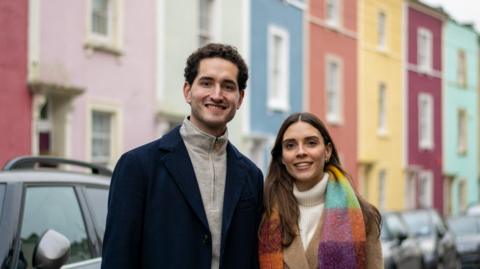 Maria Aguillon and Santiago Muriel standing in front of rainbows homes in Bristol. Maria has long brown hair and is wearing a multi-coloured scarf. Santiago is wearing a long navy coat with a grey top underneath. They are both smiling.