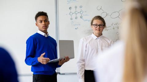 Two GCSE students presenting their findings on a physics project, holding a laptop at the front of a classroom.
