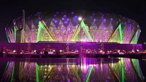 The outside of a round stadium with green and purple lights on it in the dark.