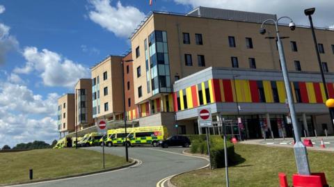 The outside of the Royal Stoke University Hospital, which is partly yellow, red and black. There are ambulances outside the building, on the left and in the middle of the image.