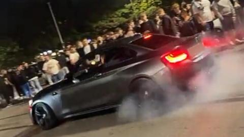 Footage from the car meet of a black car skidding its tyres in front of a large crowd.