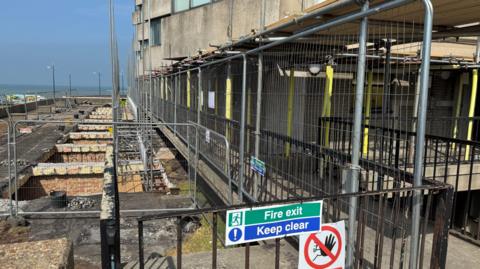 Metal fencing, scaffolding and temporary supports surround repair works outside a seafront tower block. Excavated sections of ground run alongside the building, with the sea visible in the distance and a sign reading "Fire exit Keep clear" attached to a gate.