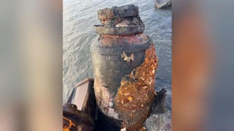 The ordnance out of the water, there is sea in the background and it is in the clasp of the digger. It is a large, dark grey colour with signs of erosion and rusty colour rocks stuck to the right hand side of it