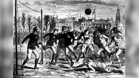 A black-and-white illustration shows an early football match with players crowding around a goal area during active play.
Multiple players in long-sleeve kits and boots move toward a ball suspended in the air, while one player lies on the ground in the foreground.
Spectators, buildings, and a clock tower in the background suggest a historic urban setting and an early era of the sport.