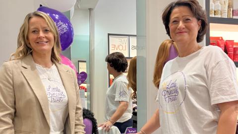 Two women stand in a hair salon. Both are wearing white T-shirts with the text Little Princess Trust. The woman on the left is wearing a beige blazer. A large mirror can be seen behind them.