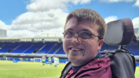 Harrison Mayhew-Kemp smiles at the camera while sitting in his wheelchair on the edge of the Portman Road football pitch. Players can be seen behind him on the pitch. He has short brown hair that hands over his forehead. He wears glasses and a burgundy top. 