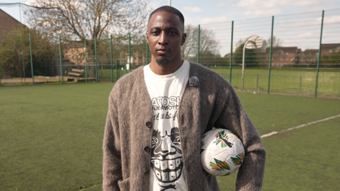 Joe Aribo stands with a football tucked under his left arm and looks directly into the camera while wearing a fuzzy fawn-coloured cardigan and white t-shirt with a black cartoon face on it. Aribo is standing on an astro turf pitch surrounded by a high green chain-link fence. Beyond the fence a park can be seen with blocks of flats beyond that