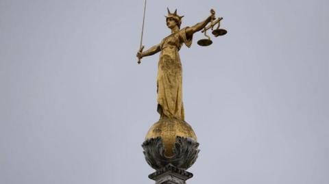  A statue of lady justice holding the scales of justice. The gold statue is on top of a stone pillar and curved holder. 