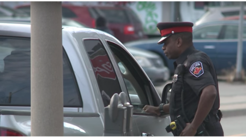 A police officer approaches the man who yelled an obscenity at a reporter from his vehicle