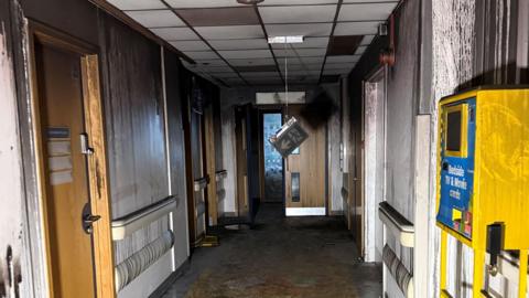 A hallway in a hospital can be seen with signs of fire damage. A fire exit sign hangs on its wire from the ceiling. Elsewhere, dirt and patches of black can be seen on the floors and walls. 