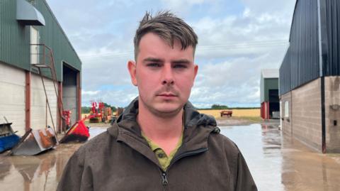 Harvey is looking into the camera with a serious expression. He's wearing a brown waterproof jacket with a green button up shirt underneath. Behind him, in the distance, is a field of yellow crops, with some red farm machinery on the edge of the field. Closer to the camera, on either side of the image, are two large barns - one is clad in green corrugated iron, and the other in black corrugated iron. The sky is cloudy with a few spots of blue visible. It's clearly rained recently as the concrete farmyard has lots of puddles. 
