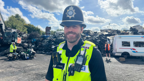 Sgt David Eeles pictured smiling while stood in a scrap yard. He is wearing a black police helmet and police yellow jacket