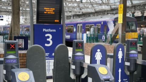 Ticket barriers at a train station, with a train in the background, and electronic departure boards displaying information