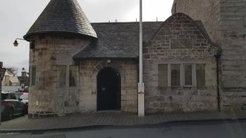 The exterior of the former police station in Castletown. It is a brown stone building with a cone-shaped roof on one side. The building is located on a street corner.