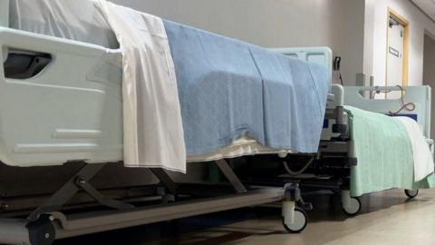 Two empty hospital beds in a corridor in a hospital corridor. The have white sheets and one has a pale blue blanket while the other is pale green.