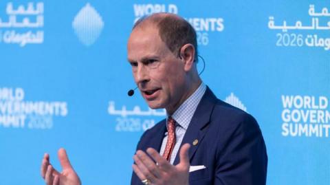 Prince Edward speaks at World Governments Summit in Dubai. He wears a blue suit jacket, blue and white checked shirt and a red tie. His hands are raised to chest height as he gesticulates whilst speaking. He sits in front of a blue board with 'World Governments Summit' printed in white behind him.