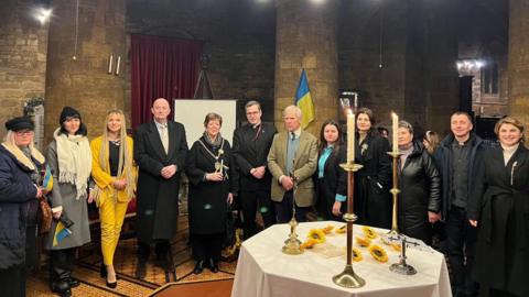 A large group of people standing inside a church, behind a table with a white table cloth on it, with candle sticks, lit, on it. There is a Ukraine flag by a wall. they are all looking at the camera. The church is made of large stone.