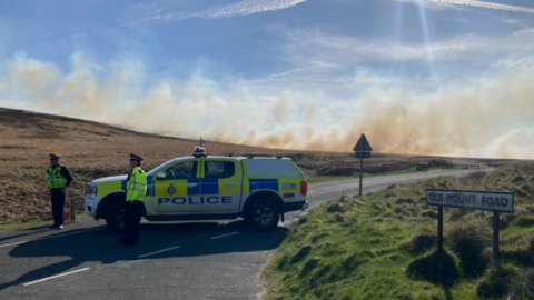 Side view of a West Yorkshire Police vehicle parked across a moorland road with two officers standing beside it. In the background a plume of brown and white smoke drifts across the moorland partially obscuring n otherwise blue sky. 