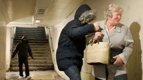 An older woman is pushed against a wall in a subway as a youth looks as if he is about to punch her on the shoulder. She has a bag and a grey coat on with grey hair. The youth has his big coat done up with a fur-lined hood over his face. Another youth also with his face covered and dark coat on and jean stands a few feet behind them. Stairs can be seen behind him.