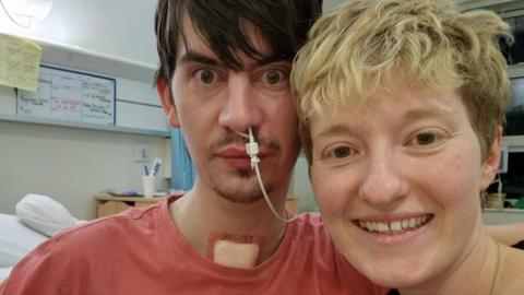 A man and woman look at the camera. The woman, with short blonde hair is smiling, the man, with dark hair, has a medical tube up his nose and a large pad on his throat