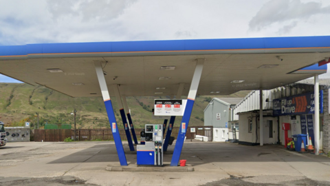 A fuel forecourt with a white and blue canopy propped up in the centre by six diagonal supports. There is a building with a sign saying Fill up & Drive to the right and a green hill in the background. 