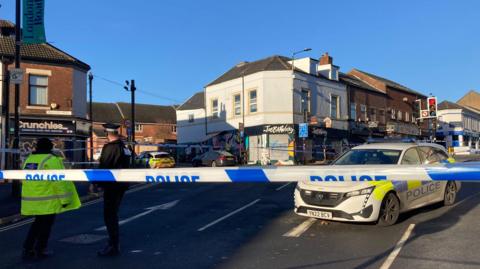 A line of police tape stretches across a road. Two police officers are standing behind the tape in the road and there are two police cars parked inside the cordon. A row of two storey buildings with shops on the ground floor. A traffic light on red stands within the cordon.