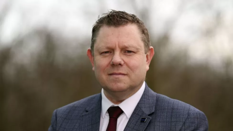 John Apter, who has short dark hair, wearing a suit, including a grey jacket and red tie