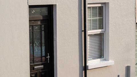 A terraced house in limewood street in Derry. On the right is a window of the home, the glass of which has been smashed when a pipe bomb was thrown at it. To the left of the window is the glass fronted front door of the property