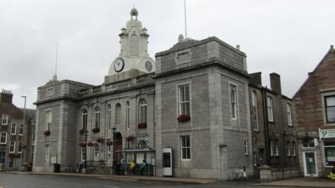 The town hall in Inverurie, a grand granite building with a bust stop in front of it and clock tower on the top and two flag poles