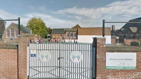 A brick wall around Avenue Junior School, Norwich, with a gate, a sign saying Avenue Junior School to the right of the gate, and beyond the gate single-storey school buildings with children playing in the distance