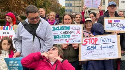 A group of people holding placards reading things like 'stop failing sen families' and a girl in a wheelchair in the foreground looking up at an adult