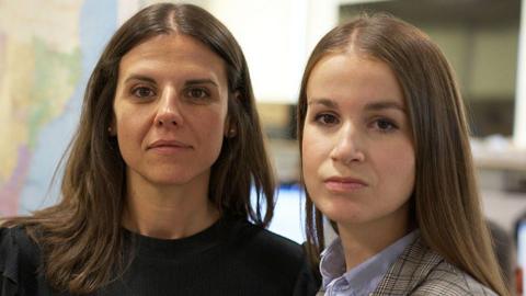 Spanish elite policewomen Cristina and Lidia look at the camera. Cristina is wearing a black top and has long brown hair. Lidia also has long brown hair and is wearing a blue shirt and checked jacket.