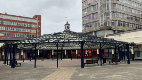 Anglia Square, a view of concrete paving, a central glass covered seating area and 1960s-style multi-storey office buildings in the background.