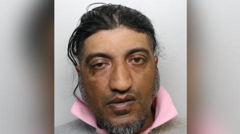 A mugshot of Nadeem Hussain who appears to have his black hair tied or combed back, he has a beard along his jaw and stubble on his upper lip. He has brown eyes with the right one appearing droopy. 