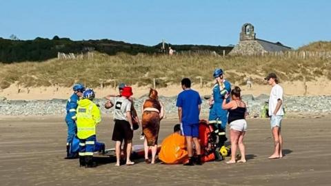 Photograph shows the back of several family members standing onshore at Llandanwg beach. Paramedics can be seen stood among them in brightly coloured blue and white uniforms.