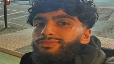 Mohammad Danyaal Asghar-Ali, 19, who has black curly hair and black beard, smiles at the camera in a close up shot taken on a residential street at night-time.