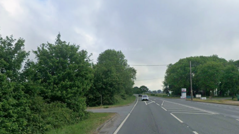Cars drive north on the A road, which is bordered by grass verges and trees. There is a white business sign in the distance.