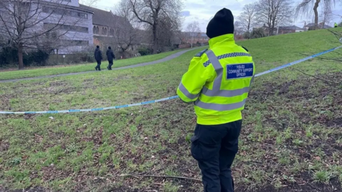 A PCSO stood facing away from the camera on a police cordon in a park, with passers-by walking past