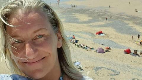 A woman with blonde hair smiling at the camera with the beach in the background. 