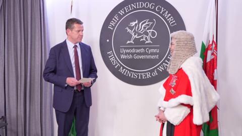 A man in a blue suit and a woman in a red and white ceremonial outfit are stood looking at one another. There is a welsh flag in the background along with white walls and a Welsh Government sign. 