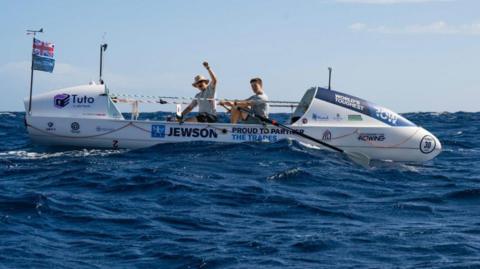 Two men on a white rowing boat on the ocean, with sponsors Jewson, Tuto and charity logo for Mind written on the side. The one at the front has his left hand raised and is grinning to the camera. He is wearing a hat and grey t-shirt. The man behind him is also looking to the camera, holding oars. He has dark brown hair.