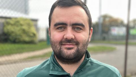 Mr Leeder wearing his Greenbank FC uniform in front of the closed pitches. 
He has a short beard and dark hair and his tracksuit is green with an emblem on his left chest.