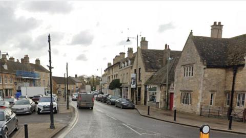 Google street view of the High Street in Market Deeping their are yellow stone buildings and shops and a car park on the left hand side