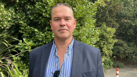 A man wearing a grey blazer over a blue and white striped shirt. He is looking into the camera. He is standing outdoors in front of a large green and bushy hedge. Sunglasses are hanging off his shirt. 