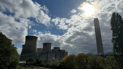 Wide view of Cottam Power Station, taken from ground level with the cooling towers and chimney rising up. Sunshine from behind clouds semi silhouettes the towers