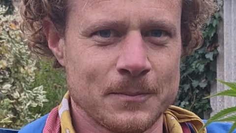 Ben Corser is smiling as he looks at the camera. He has blue eyes and light brown curly hair and a faint goatee. He is standing outdoors surrounded by plants. He is wearing a neck scarf.