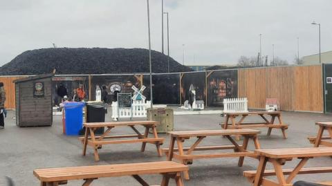 Five wooden picnic benches in the foreground, with a shed behind them, a few wheelie bins and some small models including a windmill and a white church and low white fencing are behind. Fencing panels around the mini golf have Halloween themed paintings, with one looking like a window on a castle.