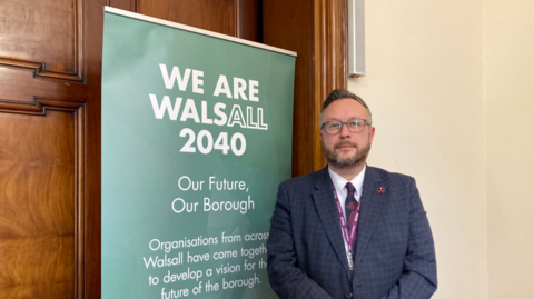 Pictured is Garry Perry with a slight smile on his face. He is wearing round dark framed glasses, has a dark blond quiff and a dark blonde beard. He is wearing a dark blue blazer with a white shirt underneath and a blue and red tie. He is standing next to a green sign that says "We are Walsall 2040".