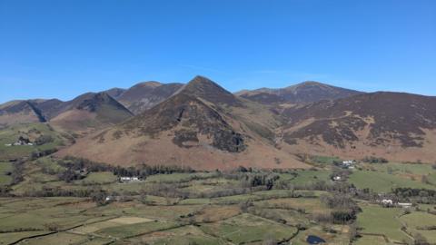 A general view of the fells taken from Cat Bells near Keswick in the Lake District. The green, undulating valley is sectioned by walls and hedges, with groupings of trees dotted around. Behind it imposing brown mountains rise up into a series of triangular peaks beneath a clear, deep blue sky.