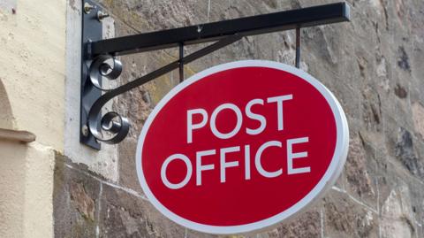 A red and white Post Office sign attached to a historic stone wall being held by a black metal beam